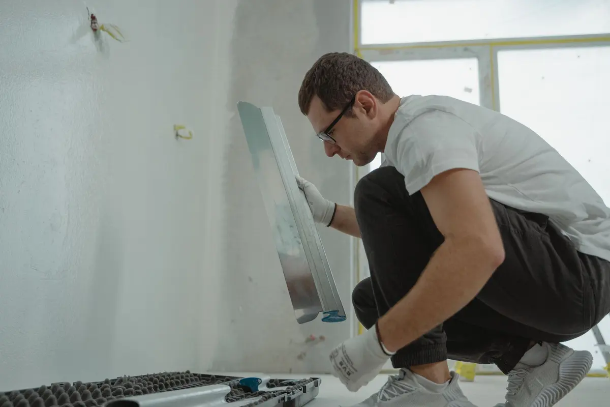 A worker kneeling to work joint compound into a wall with a wide taping knife