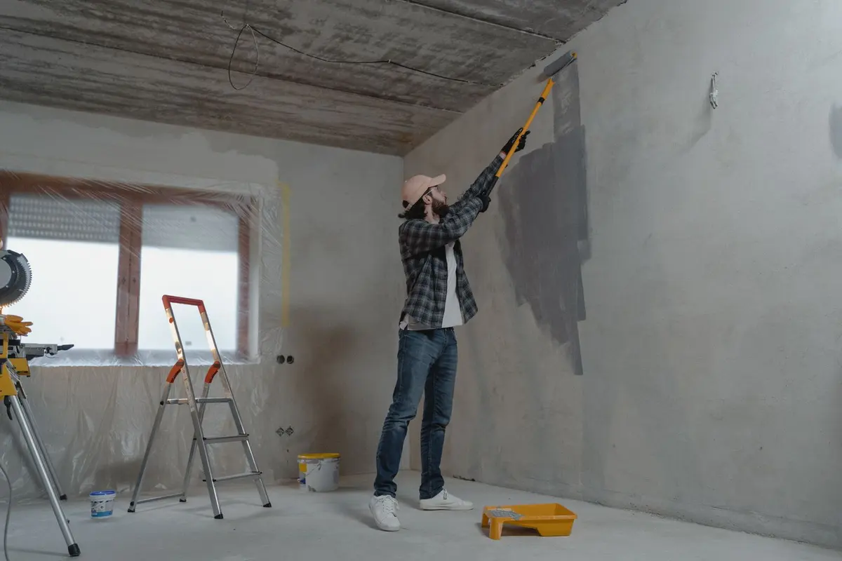 A painter rolling a fresh coat of gray paint onto an interior wall, with the floor covered and a stepladder standing by