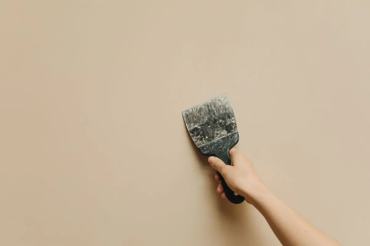 A hand holding a filling knife flat against a smooth painted wall
