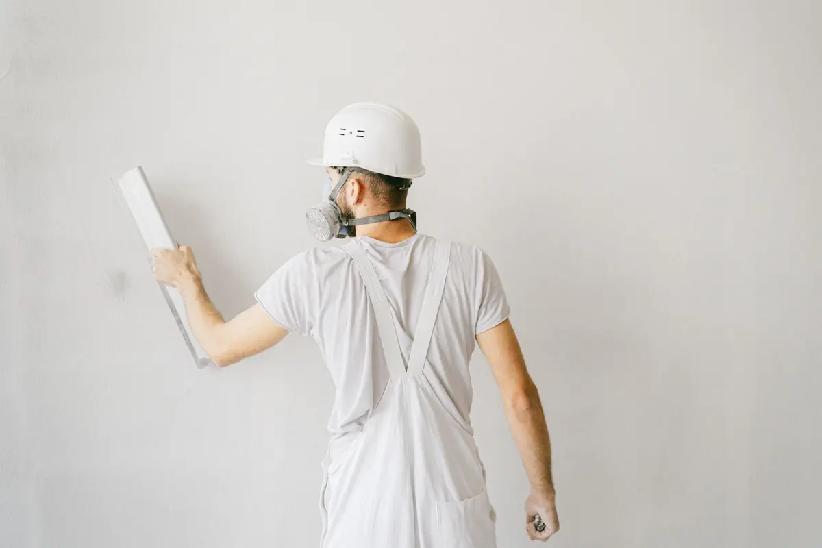 A worker in coveralls and a respirator drawing a smooth coat across a wall with a trowel